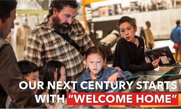 Four children and two adults gather around a touchscreen display and museum exhibit case in the Main Gallery. Text: "Our next century starts with 'Welcome Home'"