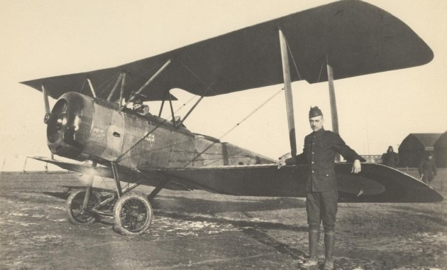 Black and white photograph of an American Soldier with a British Sopwith 1 1/2 Strutter biplane in a field in front of two buildings. There is a pilot wearing goggles in the seat and the plane's engine is running. 