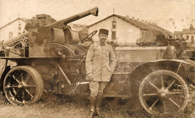 Black and white photograph of a man in a French uniform standing in front of a large piece of artillery on wheels.