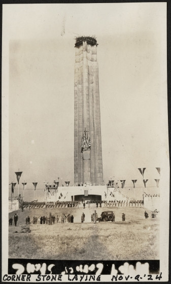 Black and white photograph of a tower under construction, with banners and celebratory decorations on it and below it. Text at the bottom reads CORNER STONE LAYING NOV-9-'24