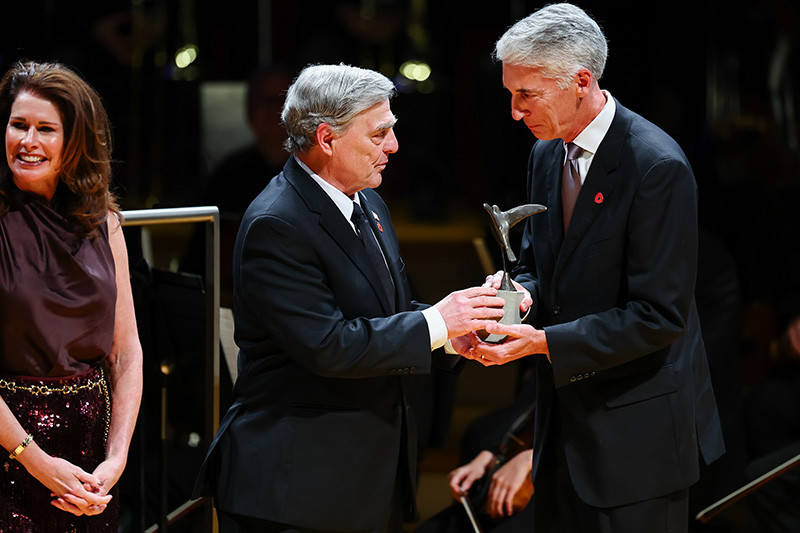 Modern photo of an older white man in a dark suit giving a dove-shaped trophy to a shorter white man also in a dark suit.