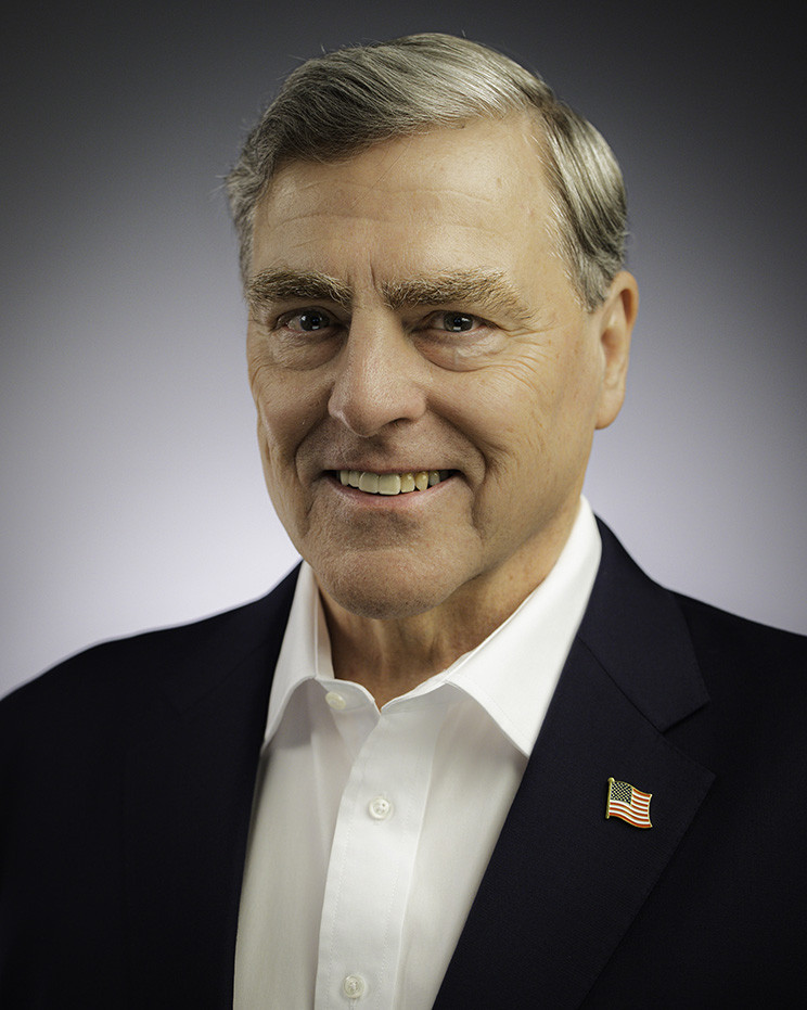 Portrait photograph of an older white man with silvered hair wearing a dark suit jacket and U.S. flag pin